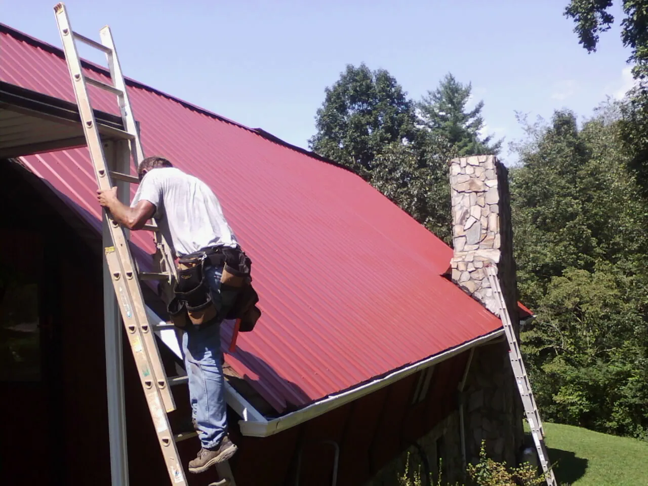 Roofer on a ladder beside a red metal roof and stone chimney — Soffit & Fascia Repair by Roofing Brothers in Olivia